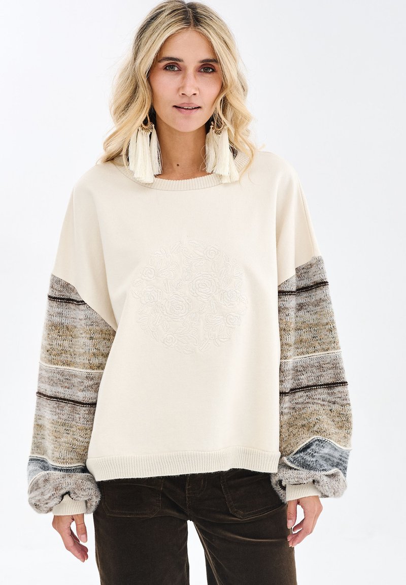 Kremowa bluza z długim rękawem o fakturowanym beżowo-szarym wzorze, z haftowanym motywem kwiatowym na przodzie i ściągaczem na dole.