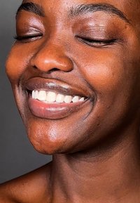 Närbild av ett leende ansikte med slät brun hud, fylliga läppar och ljusa vita tänder. Ögonen stängda, med välformade ögonbryn och en naturlig textur.