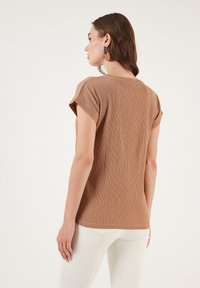 Brown textured top with short rolled sleeves and a round neckline. Worn with white pants. No visible logos or patterns.
