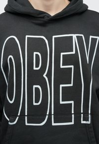 Fekete kapucnis pulóver nagy, fehér "OBEY" felirattal az elején, kenguru zsebbel a deréknál és húzózsinóros kapucnival. Puha anyagú textúra.