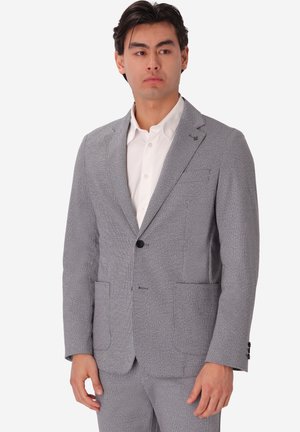Uomo che indossa una giacca da completo grigio chiaro e pantaloni abbinati sopra una camicia bianca abbottonata, in piedi davanti a uno sfondo chiaro e uniforme.