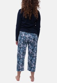 Kvinna med lockigt hår barfota, klädd i en marinblå långärmad topp och blå blommiga vida cropped byxor, stående vänd bort från kameran på vit bakgrund.