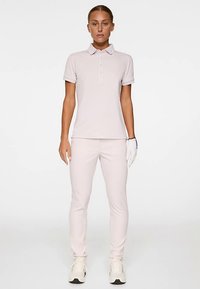 Jasnoróżowa koszulka polo z kołnierzykiem i trzema guzikami, w zestawie z dopasowanymi spodniami. Modelka ma na dłoni białą rękawiczkę golfową i sportowe buty.