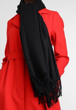 Personne portant un manteau ceinturé rouge vif et une grande écharpe noire à franges drapée autour du cou.