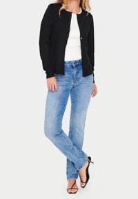 Schwarze Knopfleiste-Cardigan über einem weißen Oberteil, kombiniert mit hellblauen Jeans. Hat lange Ärmel, geraden Schnitt und schwarze Riemchensandalen.