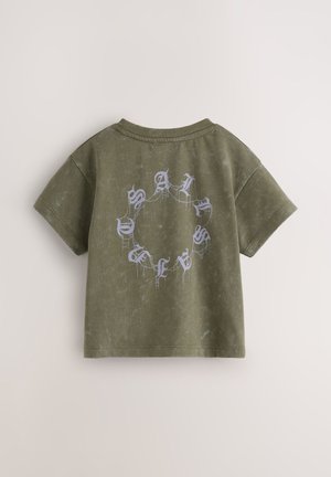 Oliwkowozielony t-shirt z krótkim rękawem i okrągłym dekoltem. Na plecach znajduje się jasnoniebieski tekst ułożony w okrągły wzór z teksturowanym wykończeniem.