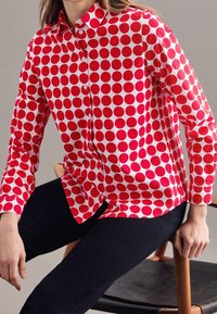 Czerwona i biała koszula w kropki z zapięciem na guziki, długimi rękawami i kołnierzykiem. Materiał wydaje się gładki z połyskującym wykończeniem.