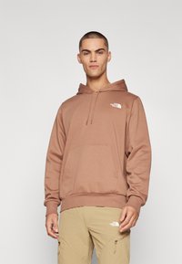 Brauner Kapuzenpullover mit einer Vordertasche, gerippten Bündchen und Saum, mit einem weißen Logo auf der Brust. Weicher Stoff und gemütlicher Schnitt.