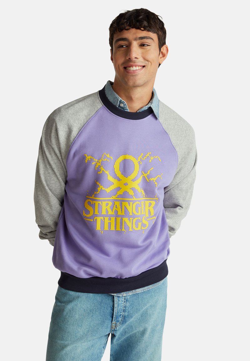 Felpa viola con maniche raglan grigie. Presenta il logo "Stranger Things" in giallo, con un motivo a fulmine. Realizzata in tessuto morbido.