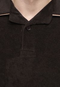 Gros plan d'une personne portant un polo texturé marron foncé avec un col boutonné et une bordure beige à l'épaule.