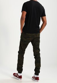 Černé triko s kulatým výstřihem, olivově zelené maskáčové tepláky s elastickými manžetami a červené boty Vans. Minimalistický design, bavlněný materiál, volný střih.