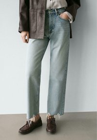 Hellblaue Jeans mit weitem Bein und ausgefranstem Saum, kombiniert mit dunkelbraunen Schnürschuhen. Darüber ein dunkler, strukturierter Blazer.