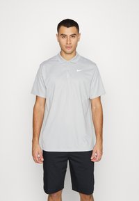Siv Nike polo majica s klasičnim ovratnikom, kratkim rukavima in belim logotipom. Ujemajoča se s temnimi kratkimi hlačami preprostega dizajna.