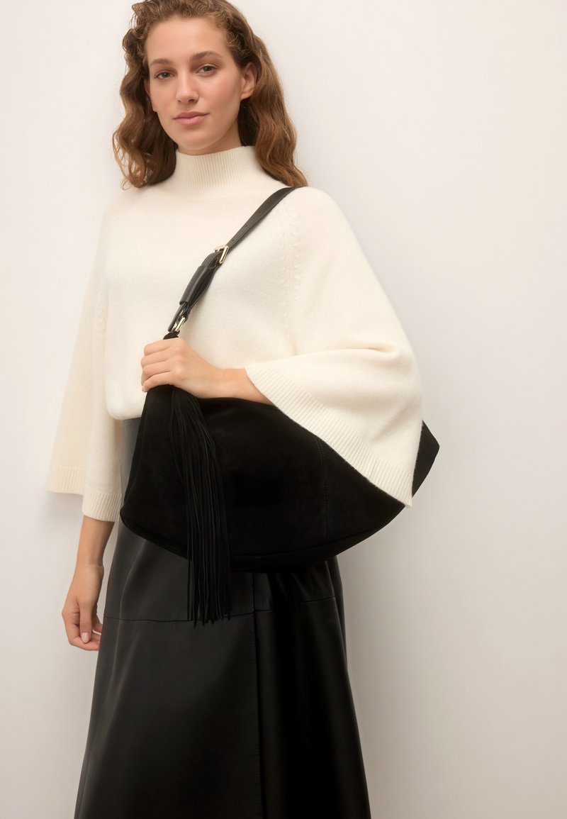 Jeune femme aux longs cheveux ondulés portant un top tricoté blanc et une jupe noire, portant un grand sac à bandoulière en daim noir avec un pompon.