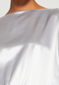 Tissu en satin argenté avec une finition brillante, présentant un large décolleté bateau et une texture lisse, tombant doucement sur l'épaule.