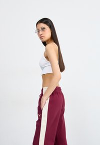 Hvid ribbet crop top parret med maroon sweatpants med en cremefarvet sidepanel og et guld logo på venstre ben. Lange mørke hår og briller.