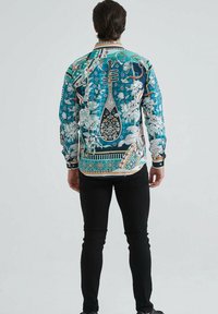 Homme de dos portant une chemise à manches longues teal avec des motifs floraux et d'instruments de musique, associée à un pantalon noir ajusté et des chaussures noires.
