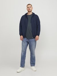 Marineblauwe zip-up hoodie met trekkoord, gedragen over een donkere t-shirt, gecombineerd met lichtblauwe jeans en witte sneakers. Glad materiaal, casual pasvorm.