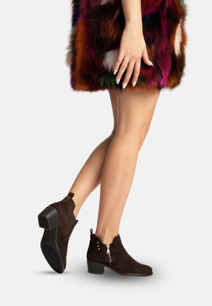 Vrouw die donkerbruine suède enkellaarsjes draagt met lage hakken en zijritsen, gecombineerd met een meerkleurige bontrok, staand op één voet.