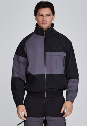 Black and gray windbreaker jacket featuring a zip front, elastic cuffs, and multiple pockets with a smooth texture and color-block design.