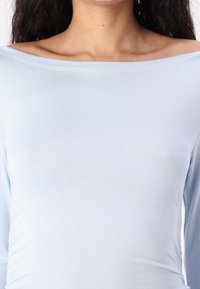 Lyseblå, langermet topp med off-the-shoulder-design. Laget av et mykt, elastisk stoff med lett rynking på siden.