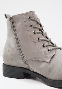 Stivale alla caviglia in suede grigio con lacci frontali, zip laterale, finitura testurizzata e tacco in gomma nera basso. Presenta occhielli metallici e dettagli di cucitura.