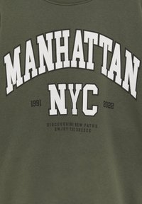 Tissu vert avec texte blanc en gras "MANHATTAN NYC" et texte plus petit "1991 2022 Discovering new paths enjoy the breeze."