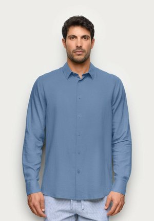 Hombre de pie frente a un fondo liso vestido con una camisa de botones de manga larga azul claro y pantalones de color claro.