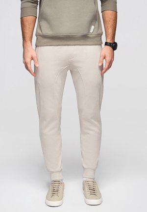 Uomo che indossa pantaloni jogger beige chiaro, sneakers abbinate, calzini bianchi e una felpa color taupe con un orologio da polso nero.