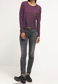 Lila Langarmoberteil mit einer Pailletten-Tasche, kombiniert mit verwitterten grauen Skinny-Jeans und schwarzen Sneakers, mit umgeschlagenen Ärmelenden.