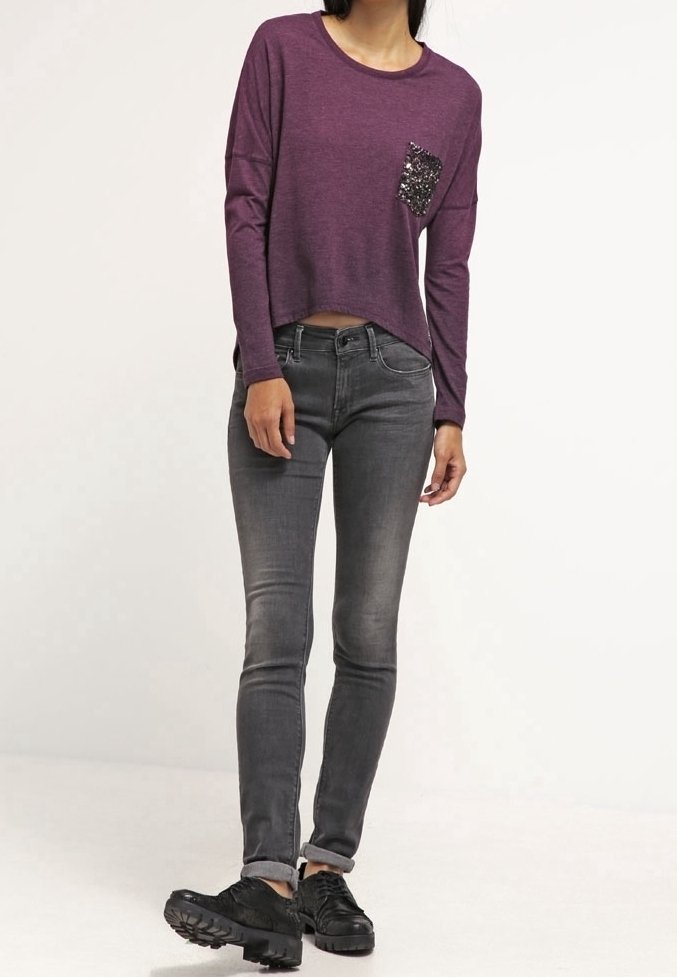 Lila Langarmoberteil mit einer Pailletten-Tasche, kombiniert mit verwitterten grauen Skinny-Jeans und schwarzen Sneakers, mit umgeschlagenen Ärmelenden.