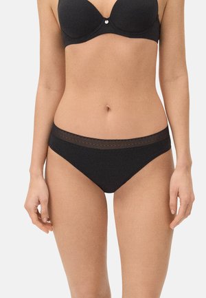 Mujer con sujetador negro y ropa interior tipo bikini negra a juego con una cinturilla estampada, de pie frente a un fondo claro y liso.