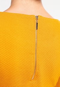 Geel textiel kledingstuk met een gouden ritssluiting aan de achterkant, met een slank ontwerp en een fijne, gemusterde oppervlakte voor extra detail.