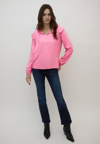 Haut à capuche rose avec épaules à volants, manches longues et cordon de serrage. Associé à un jean évasé bleu foncé et des bottines noires.