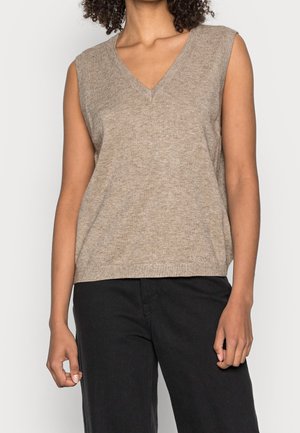 Femme portant un haut en tricot beige sans manches à encolure en V et un pantalon noir, debout devant un fond clair uni.