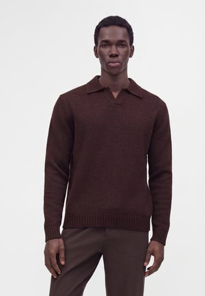 Jeune homme portant un pull col roulé marron foncé à manches longues et un pantalon marron assorti, debout devant un fond clair uni.