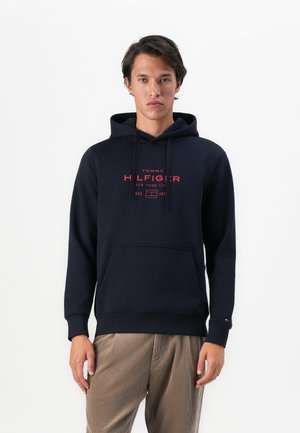 Uomo che indossa un hoodie blu navy con il logo rosso di Tommy Hilfiger, abbinato a pantaloni di velluto a coste color marrone chiaro, in piedi contro uno sfondo bianco.