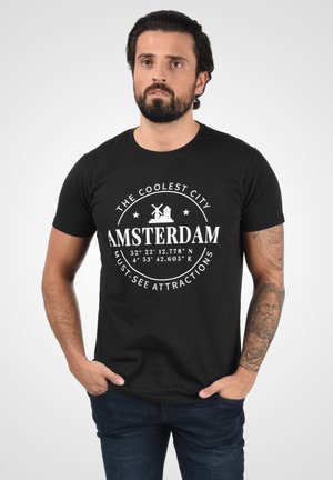 Sort bomulds t-shirt med hvidt print: "DEN SEJESTE BY AMSTERDAM" med koordinater, vindmølle og bysymboler. Korte ærmer.