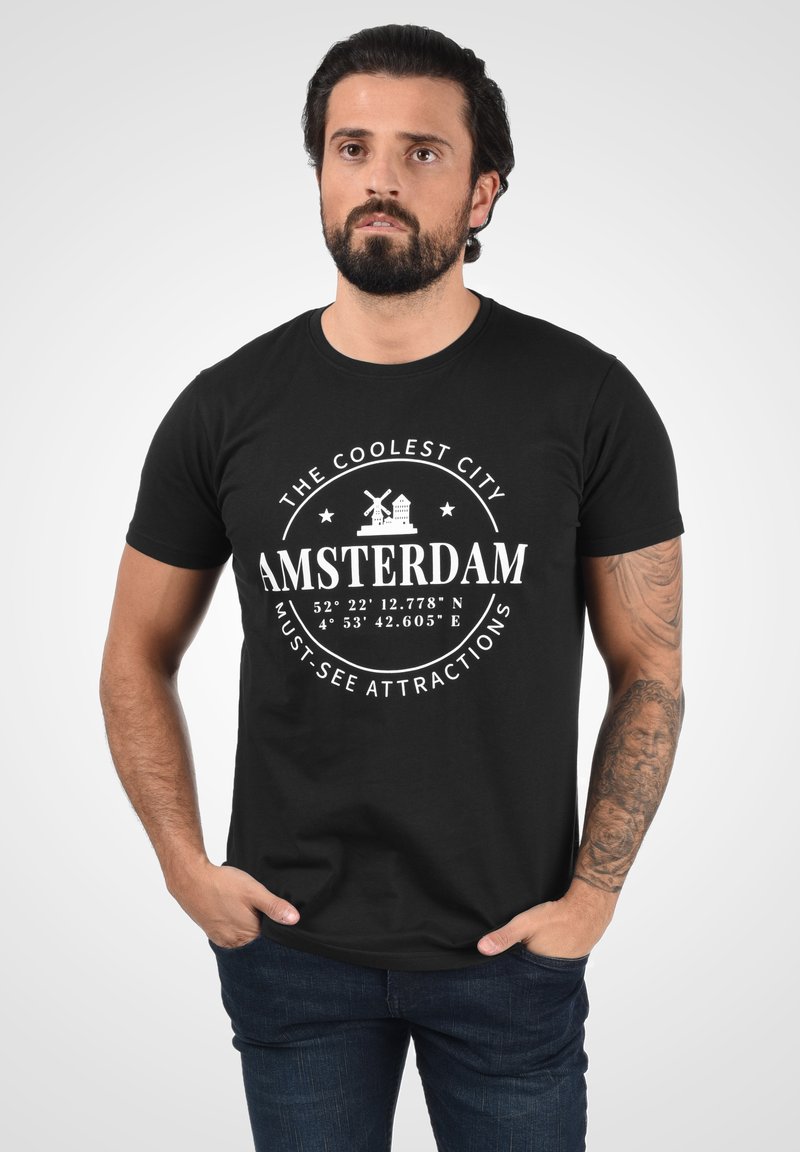 Sort bomulds t-shirt med hvidt print: "DEN SEJESTE BY AMSTERDAM" med koordinater, vindmølle og bysymboler. Korte ærmer.