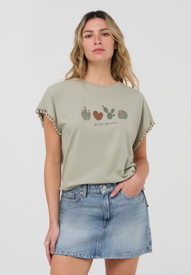 T-shirt verde oliva con bordo a pompon e grafica a tema cactus. Abbinato a una minigonna di denim blu chiaro. Design casual, texture morbida.
