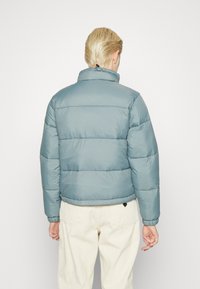 Padded jacket in light blue, featuring a high collar and elastic cuffs. Visible stitching and a cropped design. Worn with cream corduroy pants.