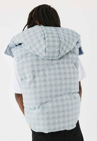 Lichtblauwe gewatteerde vest met patroon en een grote capuchon, voorzien van geometrische vormen en een gestructureerde stof, gedragen over een wit shirt.