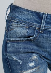 Jeans de denim azul con una textura desgastada, un ligero desvanecimiento y varios parches deshilachados. Un logo de acento está presente en el bolsillo lateral.