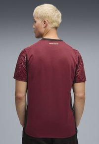 Bordeauxfarbendes Sportshirt mit kurzen Ärmeln und schwarzen Seiten, strukturierten Mustern auf den Schultern und einem runden Kragen. Goldenes Logo-Detail auf der Rückseite.