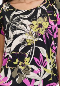 Blouse florale noire avec des motifs vibrants en rose, vert et blanc. Présente des embellissements en rhinestones transparents le long du design.