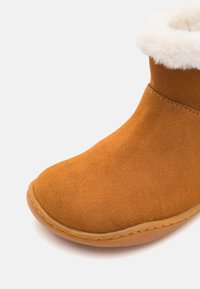 Stivaletto in suede marrone con suola marrone chiaro e imbottitura bianca e soffice visibile sul bordo superiore, su sfondo bianco.