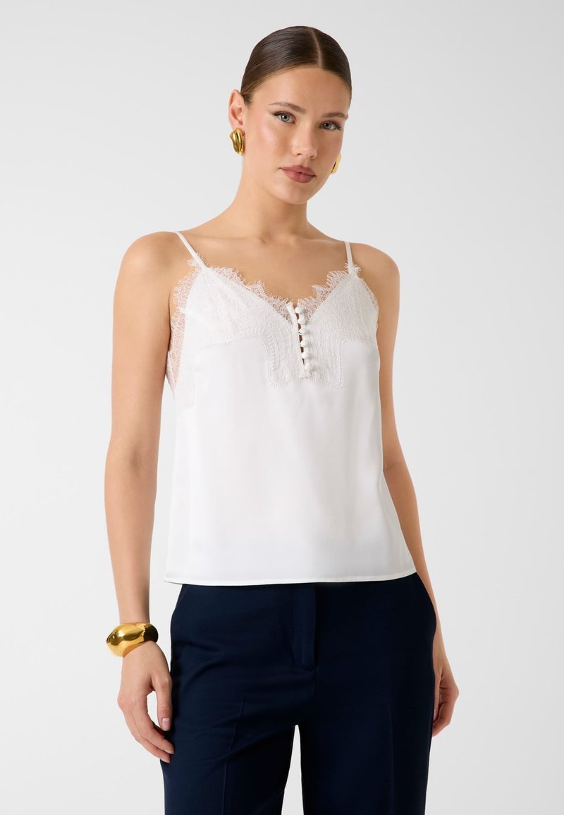 Femme portant un caraco blanc à bordure en dentelle avec des boutons, un pantalon foncé, des boucles d'oreilles en or et un bracelet en or massif, debout devant un fond uni.