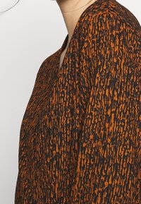 Nahaufnahme einer Person, die ein langärmliges Kleidungsstück mit einem strukturierten, orange-schwarzen, abstrakten Muster trägt.