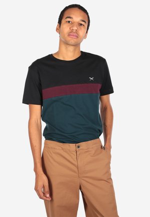 Junger Mann steht und trägt ein T-Shirt in Farbblöcken mit schwarzen, bordeauxroten und petrolfarbenen Bereichen, kombiniert mit hellbrauner Hose.