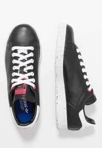 Svarta lågskor med vita snören, prickiga vita sulor och röda detaljer, visade från ovan och sidan mot en vit bakgrund.